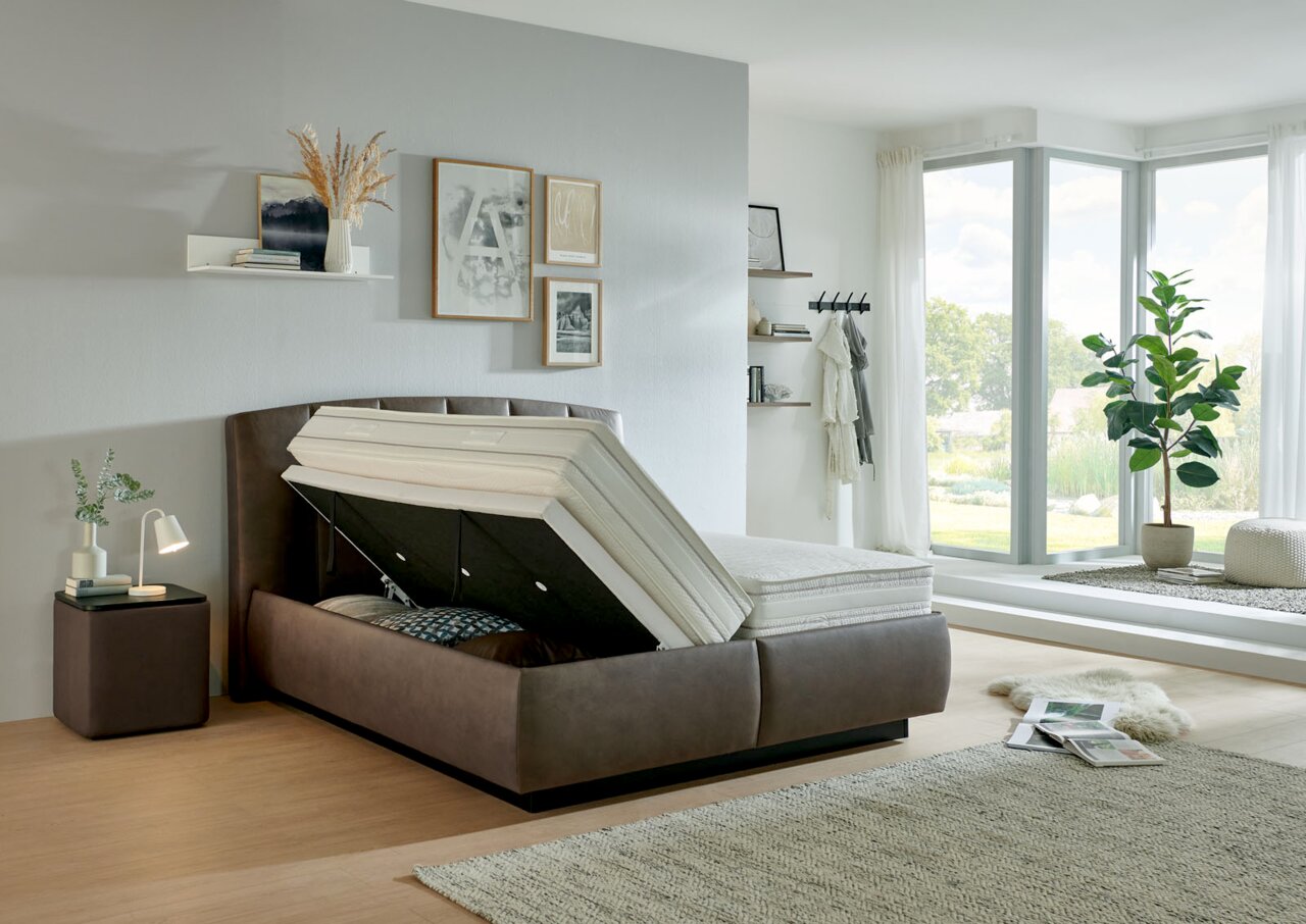 Modernes Polsterbett mit Stauraumfunktion, seitliche Perspektive, in einem hellen, stilvoll eingerichteten Schlafzimmer mit großen Fenstern.