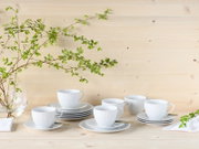 Weiße geriffelte Kaffeetassen mit Untertassen auf hellem Holztisch vor Holzpaneel-Wand, dekoriert mit grünen Zweigen und Glasvase; Perspektive: frontal auf Tischhöhe, leicht von oben.