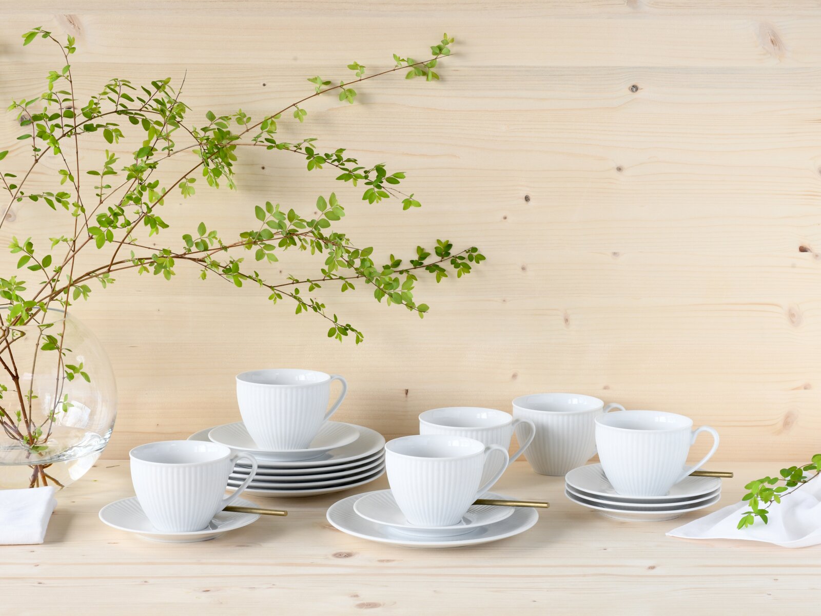 Weiße geriffelte Kaffeetassen mit Untertassen auf hellem Holztisch vor Holzpaneel-Wand, dekoriert mit grünen Zweigen und Glasvase; Perspektive: frontal auf Tischhöhe, leicht von oben.