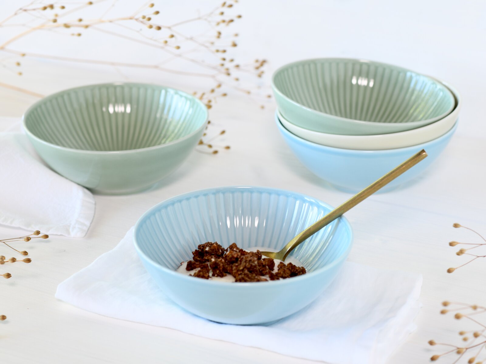 Pastellfarbene Keramikschalen mit geriffelter Innenstruktur in Mint und Hellblau, eine Schale mit Müsli und goldenem Löffel, perspektivisch in leichter Aufsicht auf weißem Tisch.
