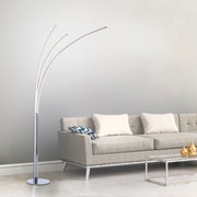 Moderne Stehleuchte aus Chrom mit drei geschwungenen Armen, seitliche Perspektive neben einem grauen Sofa.