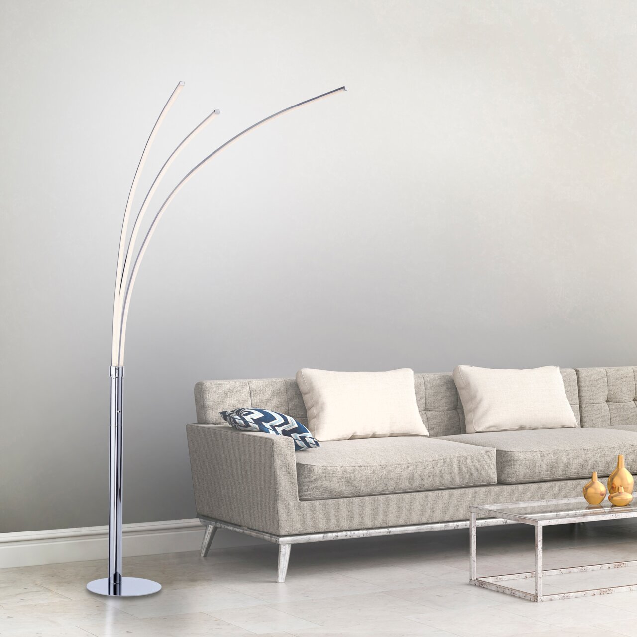 Moderne Stehleuchte aus Chrom mit drei geschwungenen Armen, seitliche Perspektive neben einem grauen Sofa.