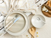 Schlichtes Geschirr-Set mit großem beigefarbenem Teller und weißer Schüssel, Besteck und Holzlöffeln auf rustikalem Tischgedeck, Draufsicht von oben