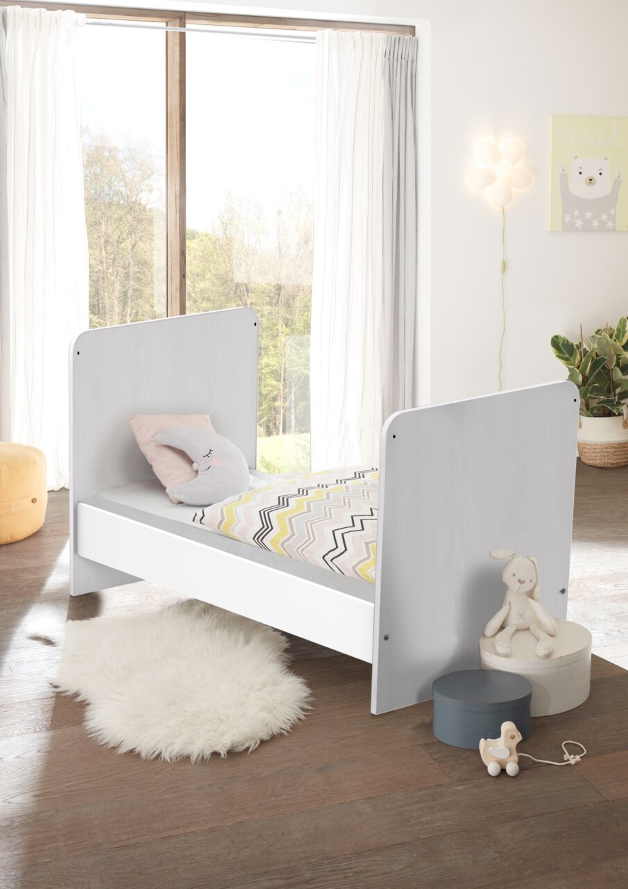 MyBaby Umbauseiten CHICO Kinderbett in einem hellen Raum mit Blick auf das Fenster, dekoriert mit einem Kissen in Mondform und einer bunten Bettdecke.