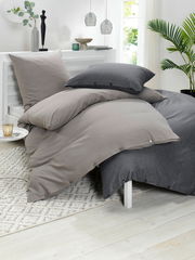 Bettwäsche-Set in Grau- und Beigetönen auf einem Bett, seitliche Perspektive, mit dekorativen Elementen im Hintergrund.