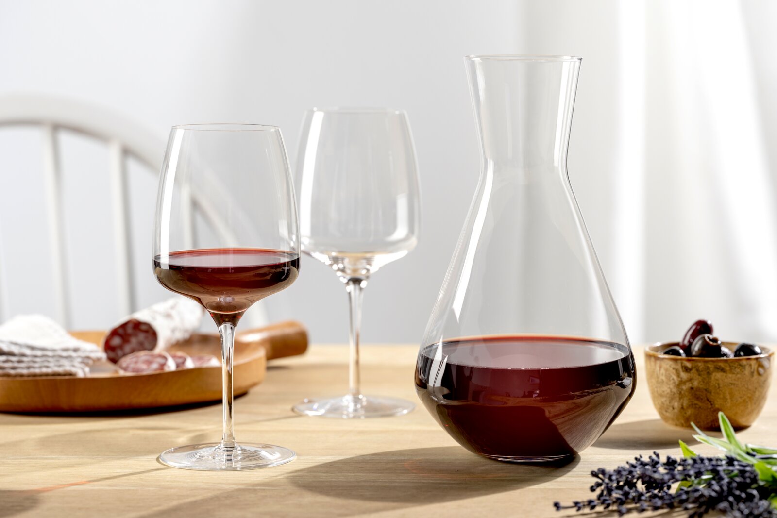 Weinset bestehend aus einer Karaffe und zwei Weingläsern, gefüllt mit Rotwein, auf einem Tisch mit einer Holzplatte und einer Schale mit Oliven, seitliche Perspektive