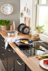 Moderne Küche mit Bratpfanne Jamie Oliver Smart auf Induktionskochfeld, seitliche Perspektive, frische Zutaten und Kräuter im Hintergrund.