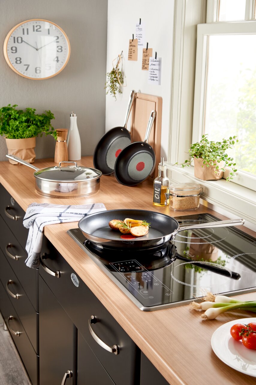 Moderne keuken met een Jamie Oliver Smart-koekenpan op een inductiekookplaat, zijaanzicht, verse ingrediënten en kruiden op de achtergrond.