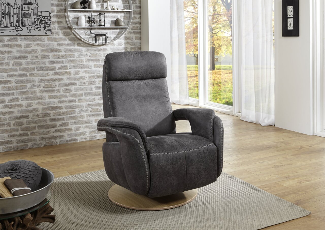 Grijze relaxfauteuil met houten onderstel, zijaanzicht, in een moderne woonkamer met grote ramen en een bakstenen muur.