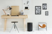 Zwarte pedaalemmer in een moderne, minimalistische ruimte met een lichte houten tafel en decoratieve wandschilderijen, gezien vanuit een zijdelingse hoek.