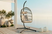 Freistehender Hängesessel in Ei-Form aus Rattan-Optik mit schwarzem Metallgestell, hellen Sitz- und Rückenkissen sowie Kopfkissen auf einer modernen Terrasse mit Glasgeländer; Perspektive: frontale leicht seitliche Ansicht auf Augenhöhe.