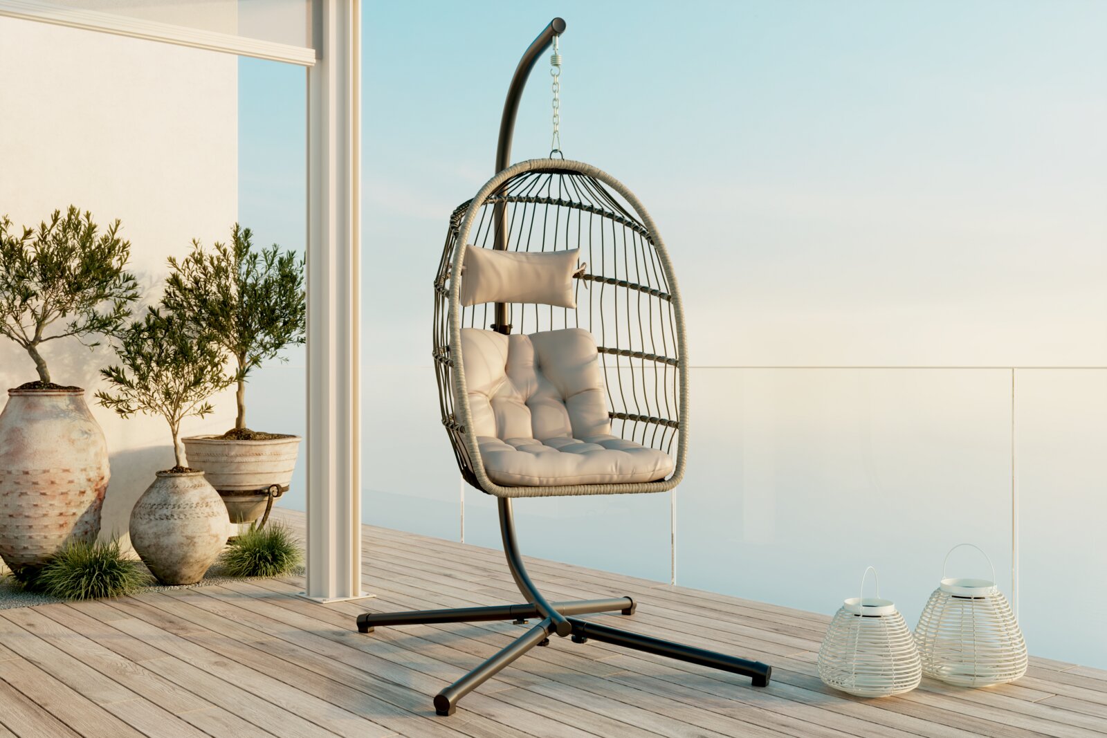 Ambia Garden Hängesessel THEO Freistehender Hängesessel in Ei-Form aus Rattan-Optik mit schwarzem Metallgestell, hellen Sitz- und Rückenkissen sowie Kopfkissen auf einer modernen Terrasse mit Glasgeländer; Perspektive: frontale leicht seitliche Ansicht auf Augenhöhe.
