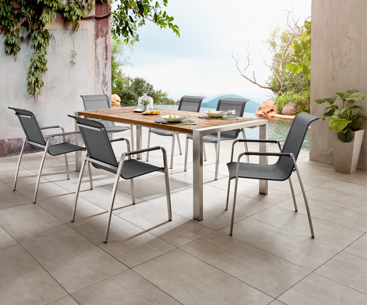 Moderner Gartentisch mit Metallrahmen und Holzplatte, umgeben von sechs stilvollen Stühlen, auf einer Terrasse mit Blick auf einen Pool und üppige Vegetation, seitliche Perspektive.