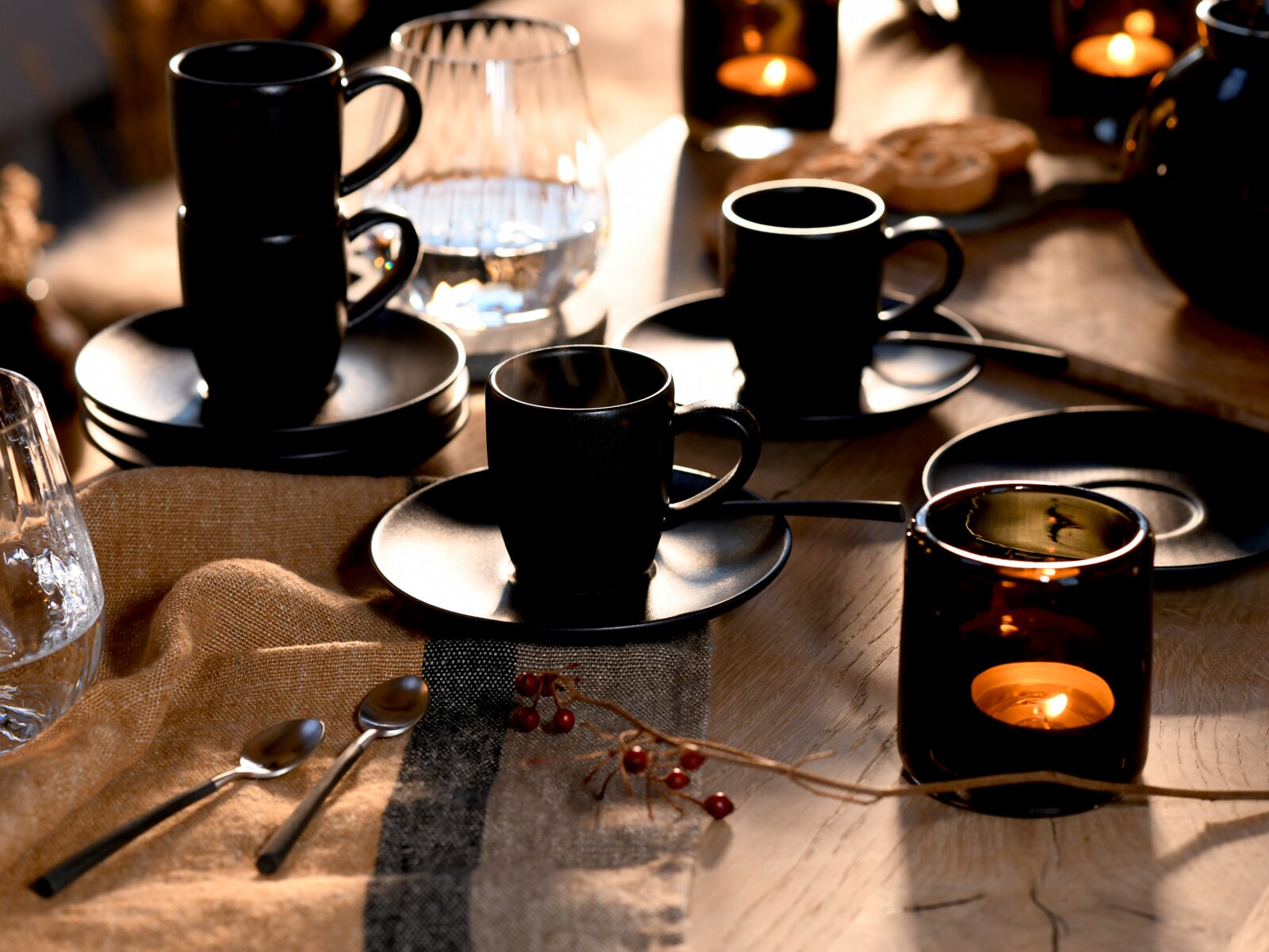 Stilvoll gedeckter Tisch mit schwarzen Espressotassen auf Untertassen und Teelichthaltern, warmes Licht; Perspektive: leicht von oben in schräger Seitenansicht.