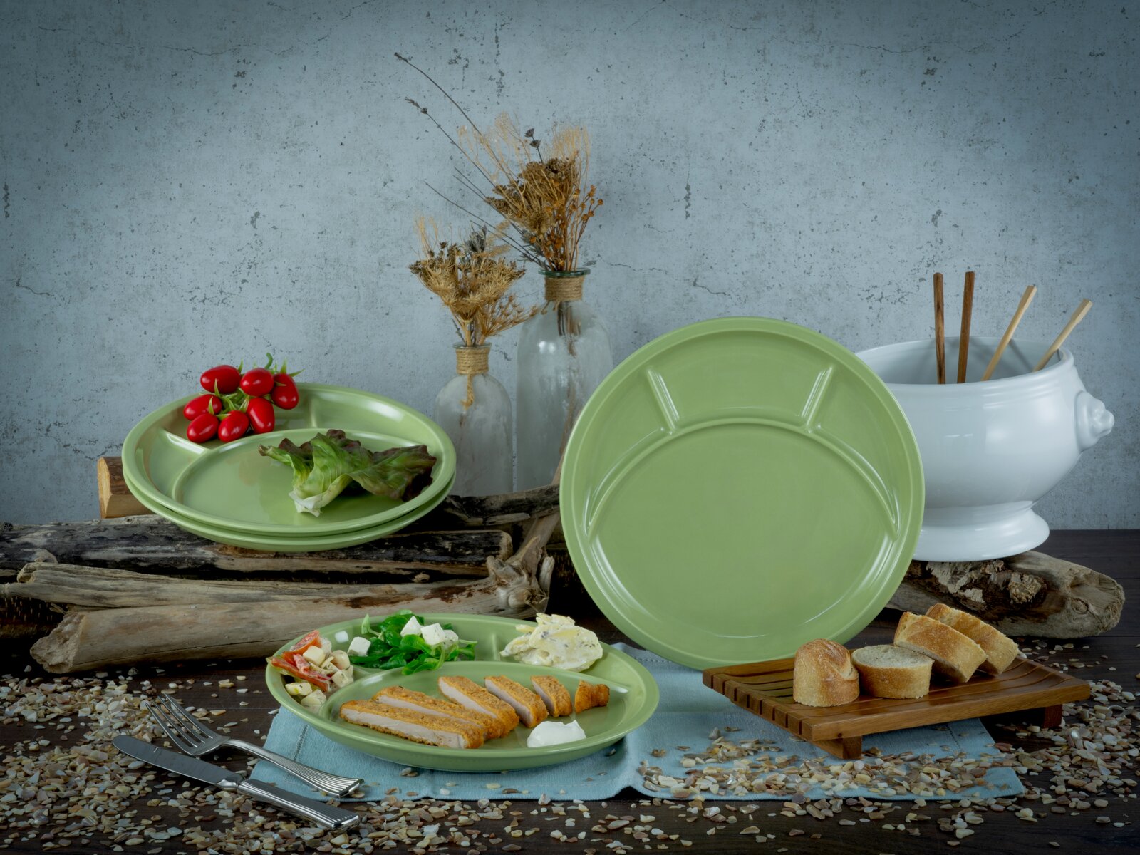 Grünes, glänzendes Servier- und Menüteller-Set mit Unterteilungen auf rustikalem Tisch, frontal in leichter Schrägansicht, dekoriert mit Salat, Tomaten, Brot und Besteck