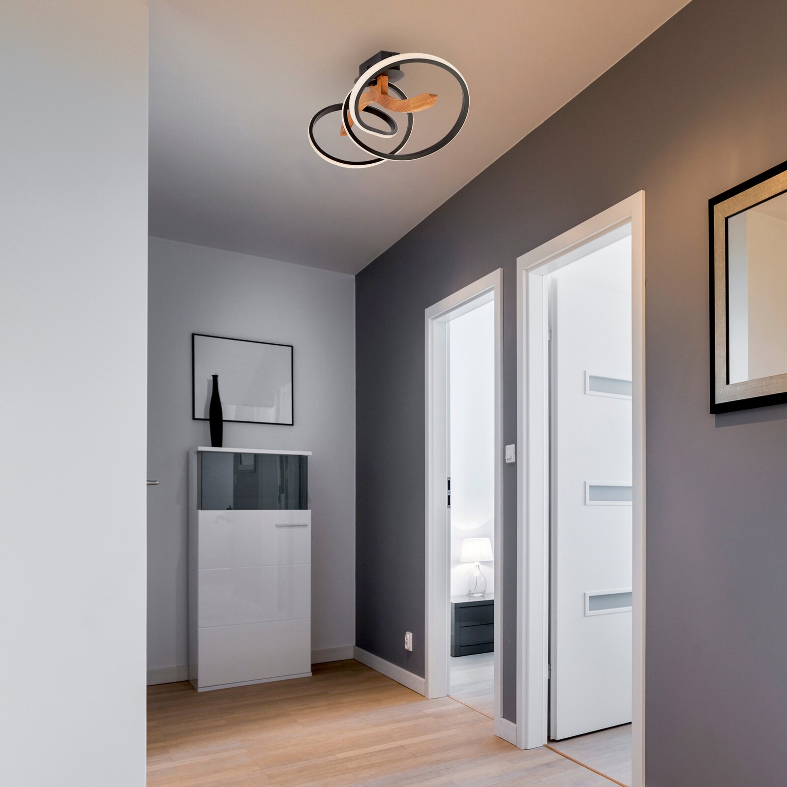 Moderne LED-Deckenleuchte mit zwei ineinander verschlungenen Ringen, von unten betrachtet in einem stilvollen Flur mit grauen Wänden und Holzboden.
