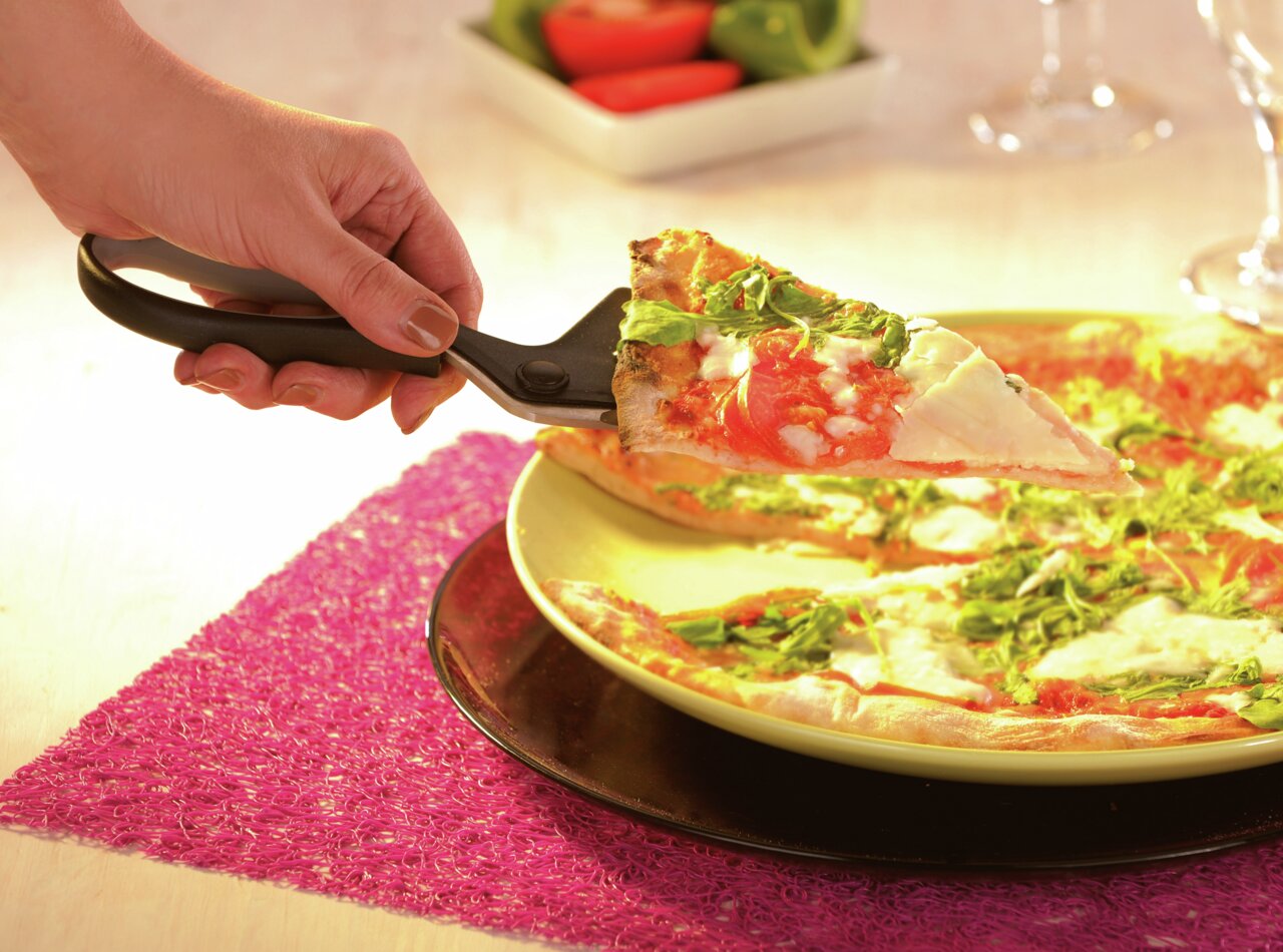 Een hand houdt een pizzaschaar vast waarmee een stuk pizza met tomaten en rucola van een bord wordt gesneden. Bovenperspectief op het bord.