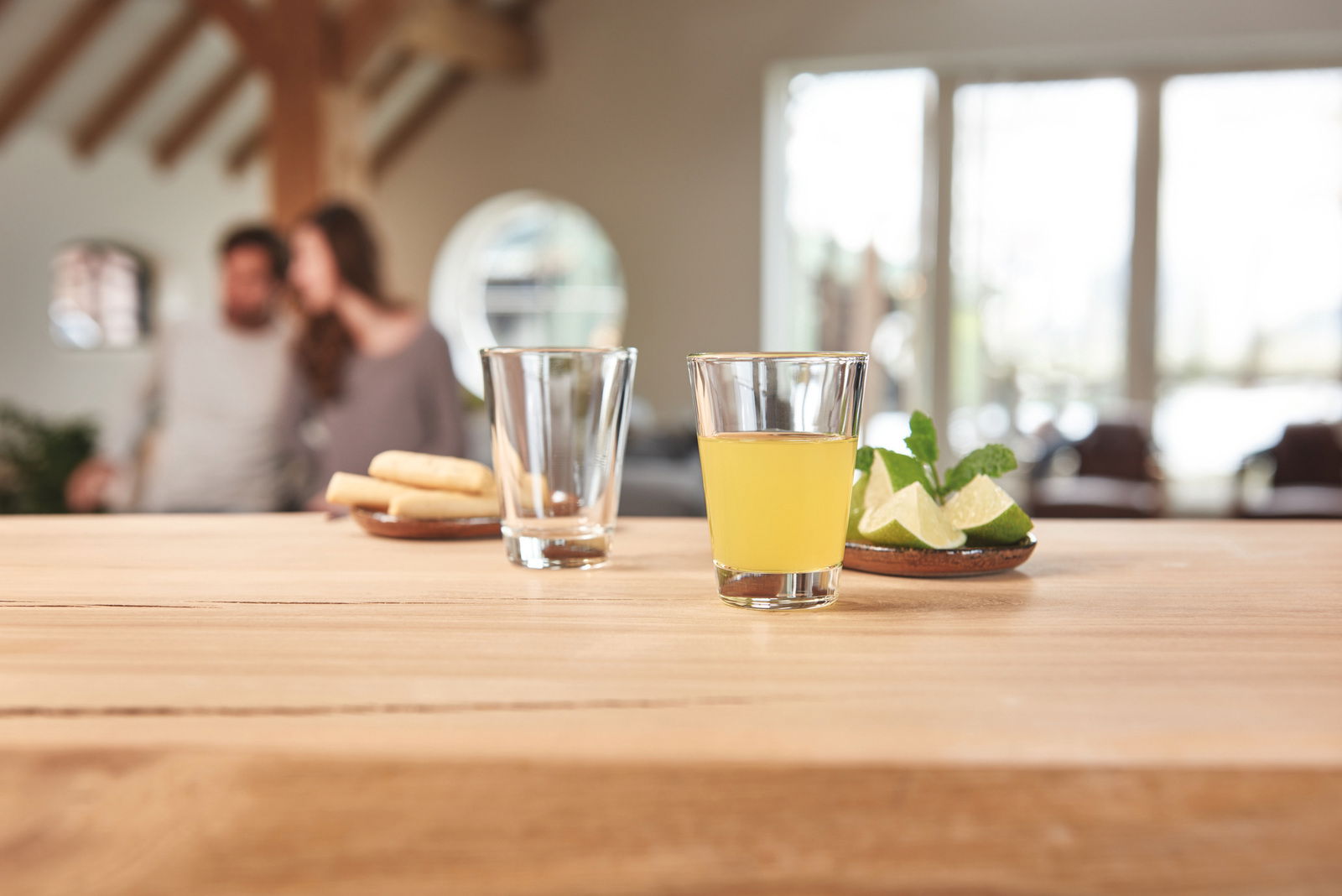 Ein Glas mit gelbem Getränk auf einem Holztisch, daneben ein leeres Glas und ein Teller mit Limettenscheiben und Minze. Im Hintergrund unscharf ein Paar in einem gemütlichen Raum. Perspektive: Nahaufnahme des Glases im Vordergrund.