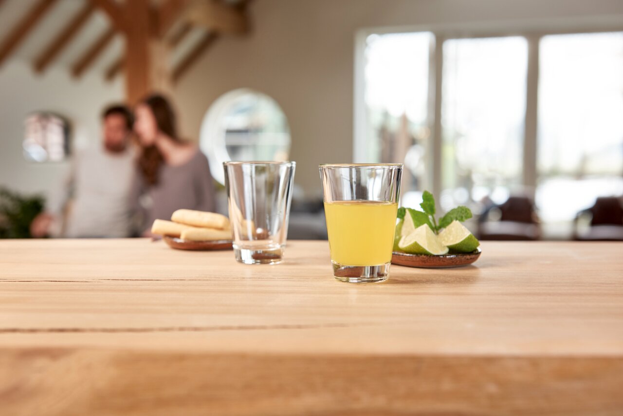 Ein Glas mit gelbem Getränk auf einem Holztisch, daneben ein leeres Glas und ein Teller mit Limettenscheiben und Minze. Im Hintergrund unscharf ein Paar in einem gemütlichen Raum. Perspektive: Nahaufnahme des Glases im Vordergrund.