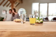 Een glas met een gele drank op een houten tafel, ernaast een leeg glas en een bord met schijfjes limoen en munt. Op de achtergrond, wazig, een stel in een gezellige kamer. Perspectief: close-up van het glas op de voorgrond.