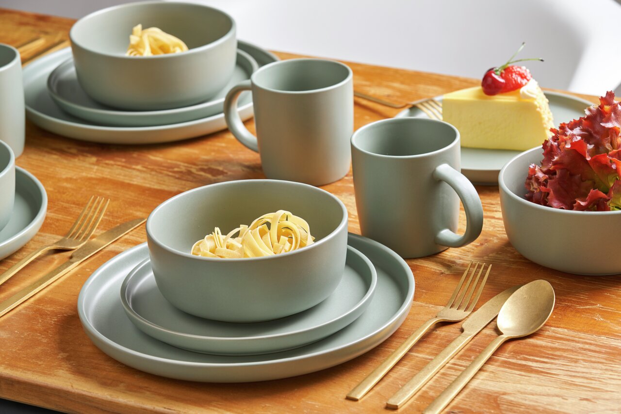 Kombiservice Prato 16-tlg. in moderner, matter Optik auf einem Holztisch, bestehend aus Tellern, Schalen und Tassen, mit goldfarbenem Besteck, aus der Perspektive von schräg oben fotografiert.