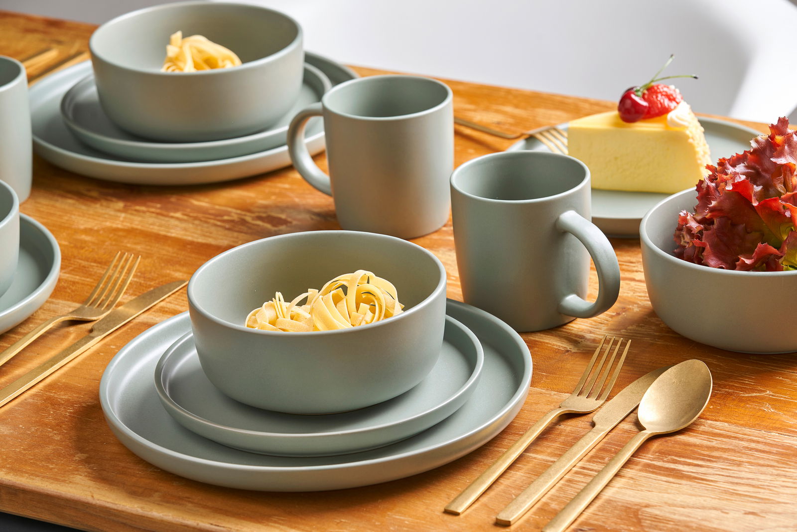 Kombiservice Prato 16-tlg. in moderner, matter Optik auf einem Holztisch, bestehend aus Tellern, Schalen und Tassen, mit goldfarbenem Besteck, aus der Perspektive von schräg oben fotografiert.