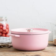 Runder rosa Gusseisen-Bräter mit Deckel, seitliche Perspektive, auf einem Holztisch mit Küchenutensilien im Hintergrund.