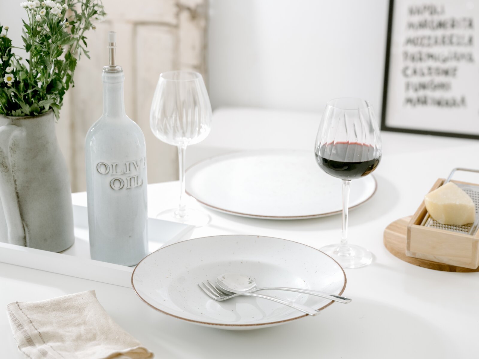 Heller, minimalistisch gedeckter Esstisch in leicht schräger Frontansicht: weißer Keramikteller mit Besteck, graue Olivenölflasche, Weingläser (eins mit Rotwein), Blumen in Vase und Käse mit Reibe im Hintergrund.