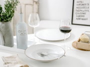 Heller, minimalistisch gedeckter Esstisch in leicht schräger Frontansicht: weißer Keramikteller mit Besteck, graue Olivenölflasche, Weingläser (eins mit Rotwein), Blumen in Vase und Käse mit Reibe im Hintergrund.