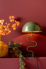 Grüne Tischleuchte mit geschwungenem Ständer und halbrundem Schirm, seitliche Perspektive, dekoriert mit Pflanzen und Vase auf rotem Hintergrund.