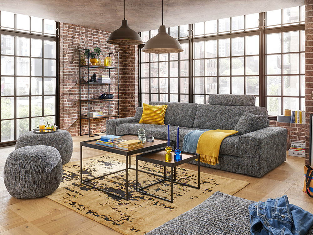 EiEin Wohnzimmer mit einem großen grauen Sofa und zwei kleinen Couchtischen.n gemütliches Wohnzimmer mit einem großen grauen Sofa und bunten Kissen. Der Raum hat eine Ziegelwand und viele Fenster, die viel Licht hereinlassen. Zwei kleine Tische stehen auf
