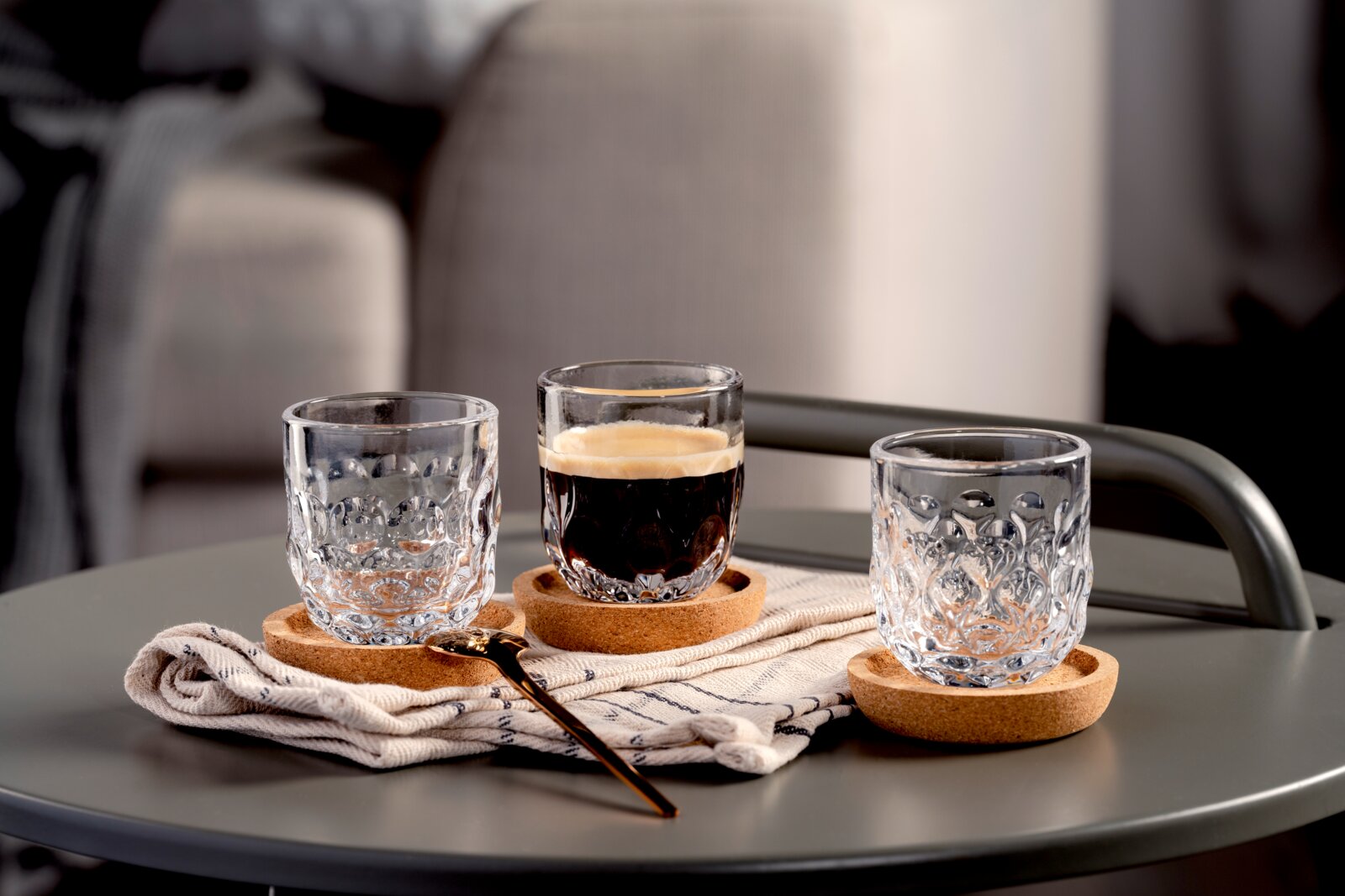 Drei elegante Espressogläser mit dekorativem Muster auf Korkuntersetzern, eines mit Espresso gefüllt, auf einem Tisch mit einem goldenen Löffel und einem Tuch, seitliche Perspektive.