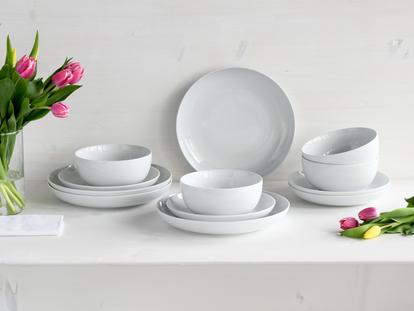 Weißes, minimalistisches Porzellan-Geschirrset mit Tellern und Schalen auf weißem Tisch, dekoriert mit pinken und gelben Tulpen; Perspektive: frontale Studioaufnahme auf Augenhöhe.