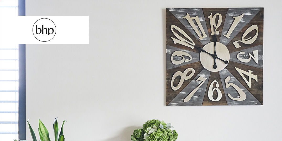 Eine große, quadratische Wanduhr mit auffälligen Zahlen hängt an einer hellen Wand.