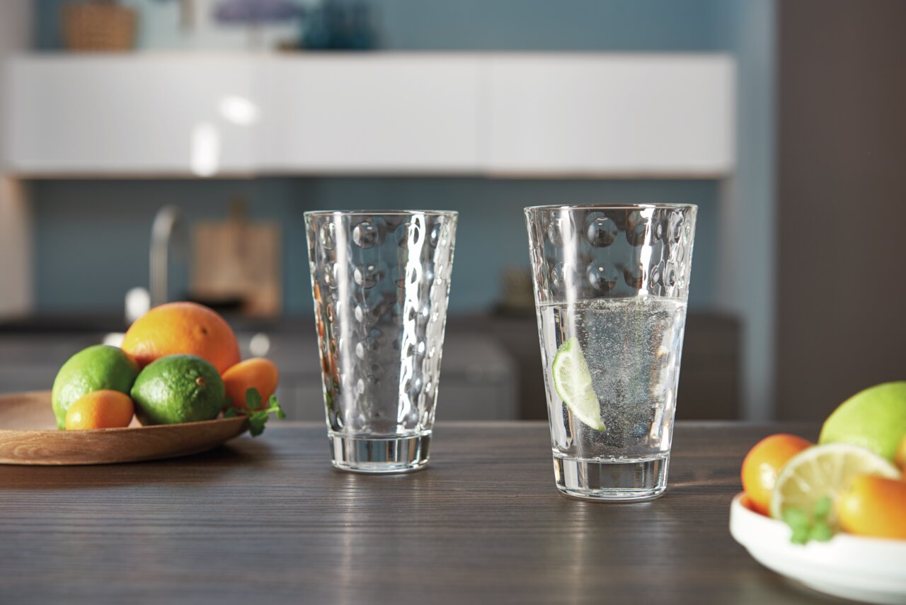 Zwei Trinkgläser Optic 300ml auf einem Tisch in einer modernen Küche, eines mit Wasser und Limettenscheibe gefüllt, im Hintergrund Obstschale mit Zitrusfrüchten, seitliche Perspektive.