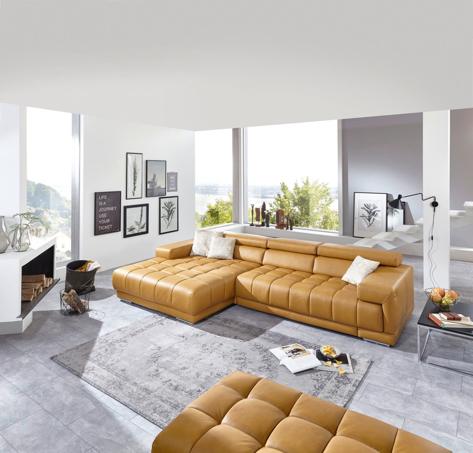 Moderne Longchairkombination in einem hellen Wohnzimmer mit großen Fenstern, die einen Blick auf die Landschaft bieten. Die Perspektive zeigt die Couch von vorne und leicht seitlich.