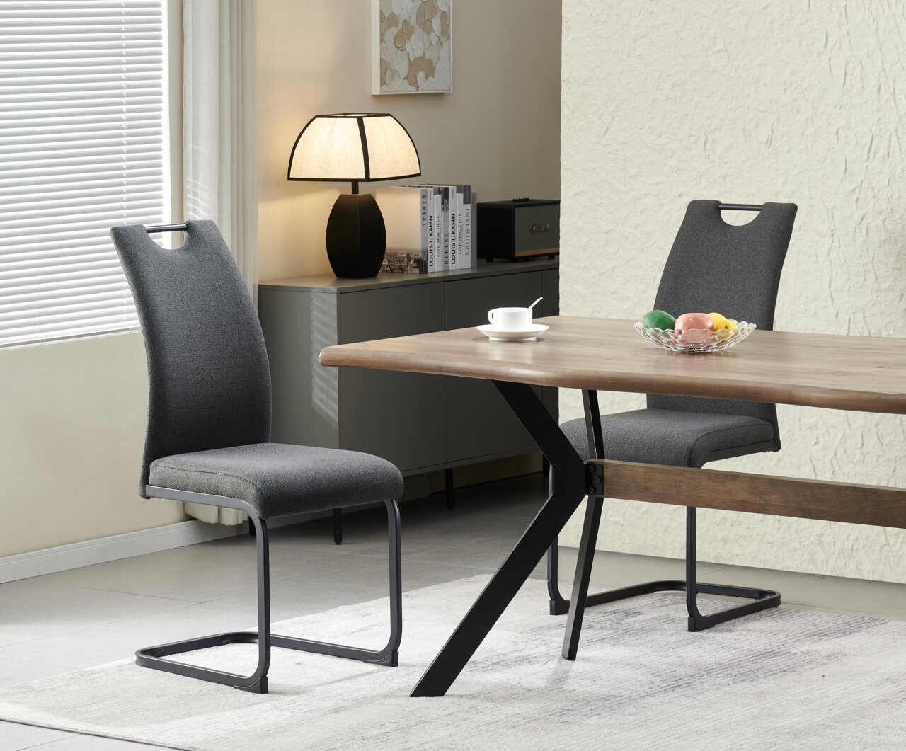MID.YOU Schwingstuhl 2er-Set HANNA Schwarzer Tischlampenfuß mit hellem Stoffschirm auf einem Sideboard neben Büchern, im Hintergrund Essbereich mit Holztisch und grauen Polsterstühlen, aufgenommen in seitlicher Perspektive.