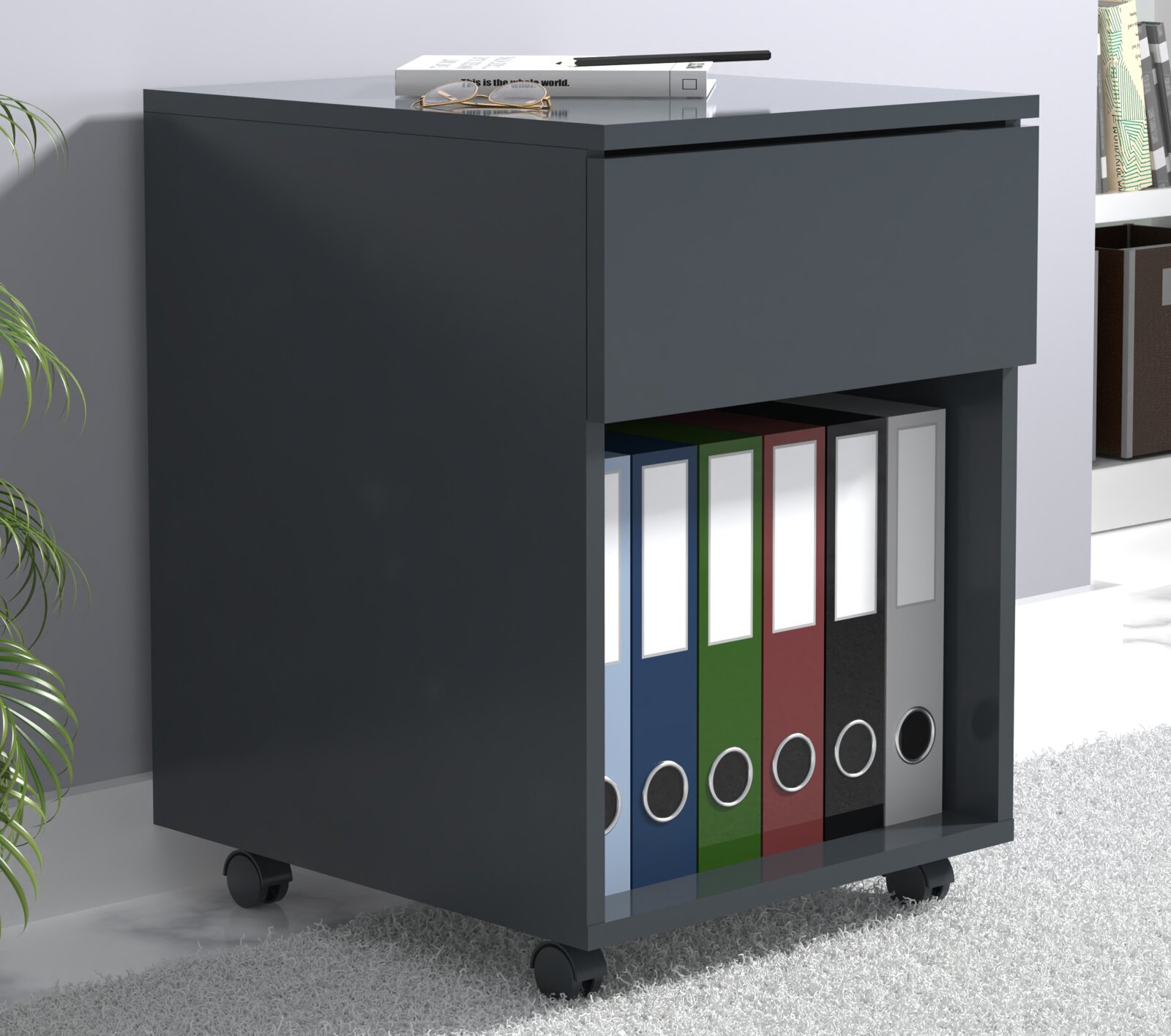 Anthrazitfarbener Rollcontainer mit Schublade und offenem Fach für Ordner, auf Rollen, in schräger Vorderansicht von links; oben liegen Bücher, Stift und Brille.