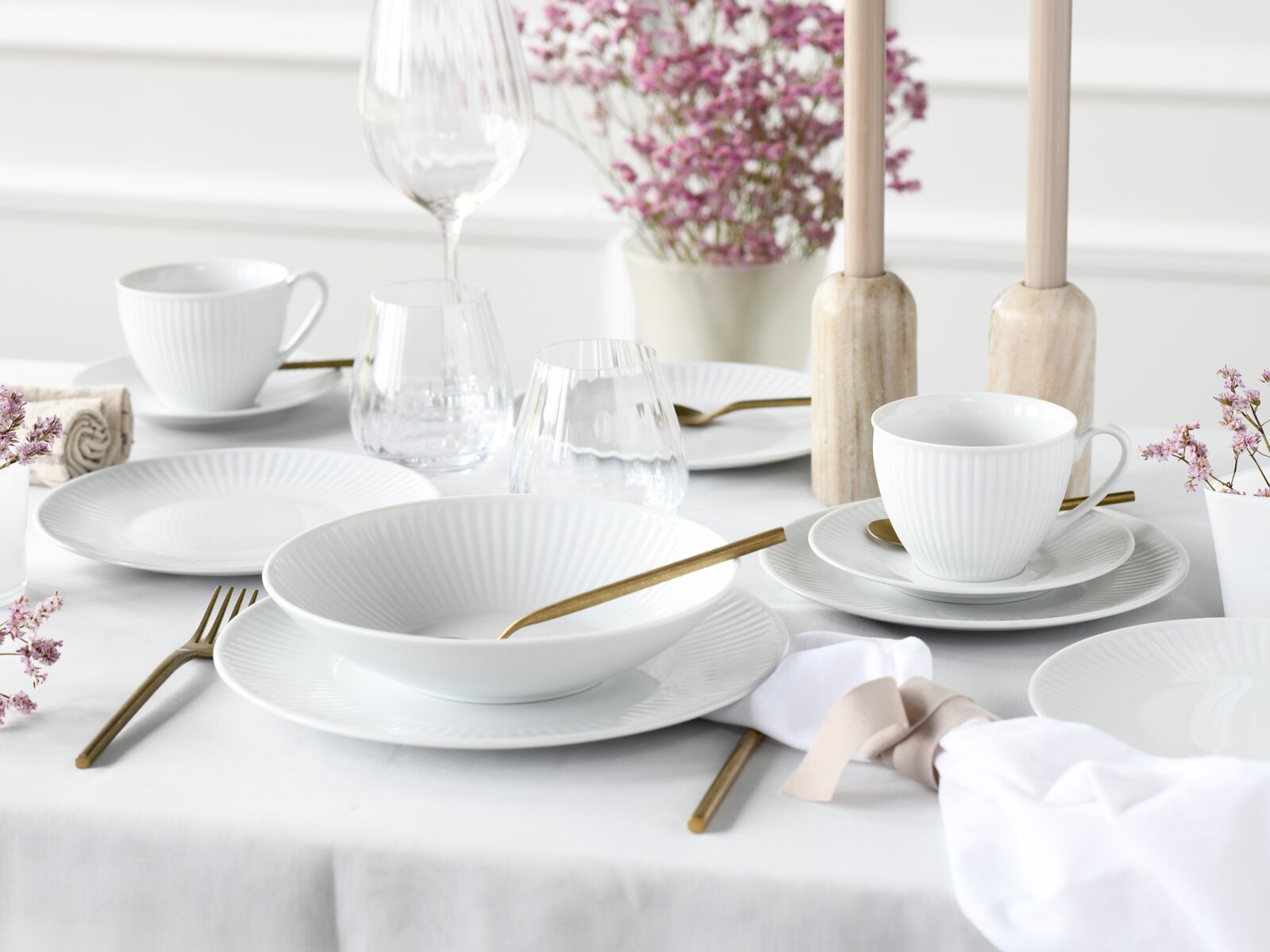 Elegant gedeckter Tisch mit weißem geripptem Porzellangeschirr (Teller, Schalen, Tassen), klaren Gläsern und goldfarbenem Besteck, dekoriert mit Kerzenständern und rosa Blumen; Perspektive: leicht erhöht aus schräger Frontansicht.