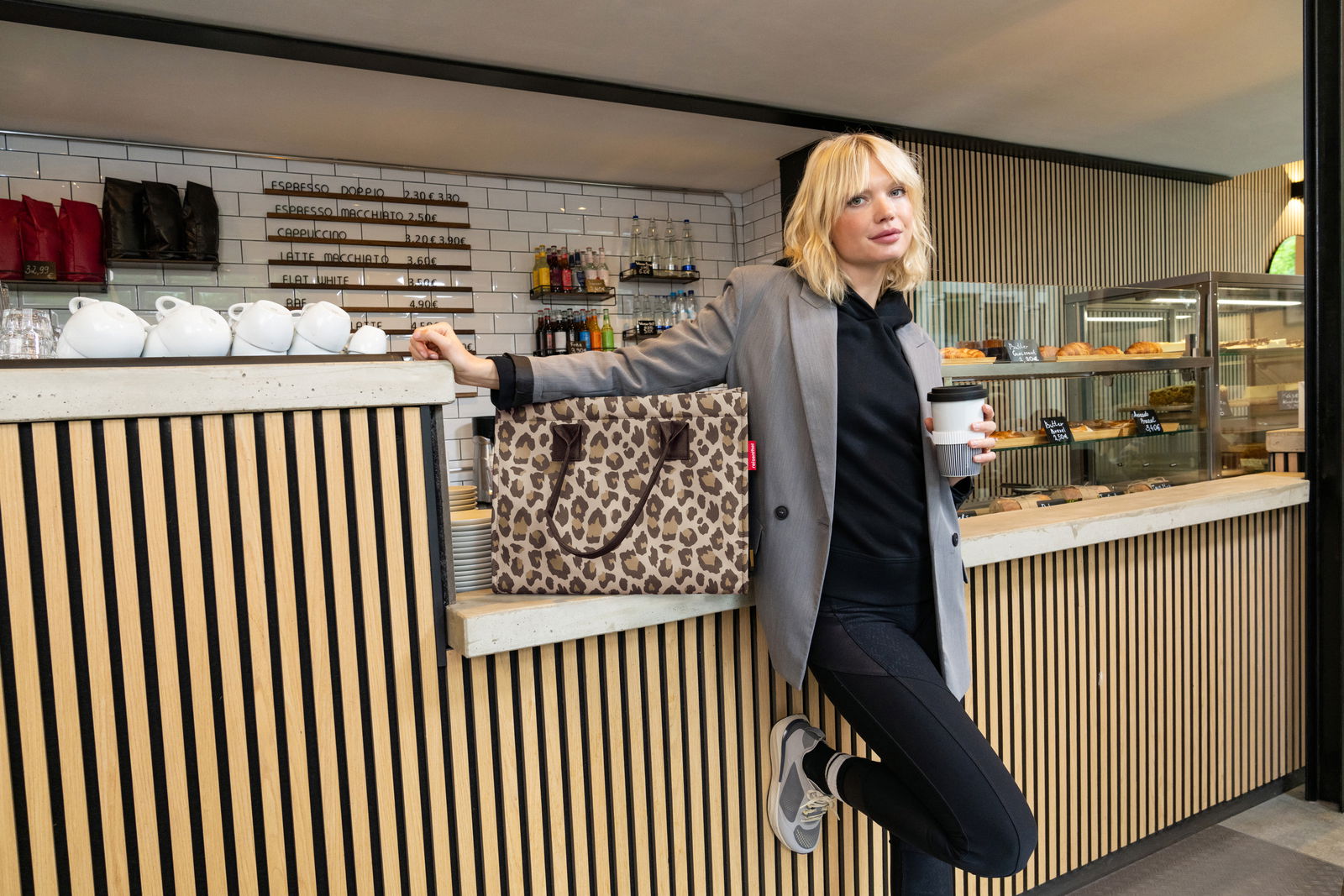 Frau lehnt an einer Theke in einem Café, hält einen Kaffeebecher und eine Tasche mit Leopardenmuster, seitliche Perspektive.