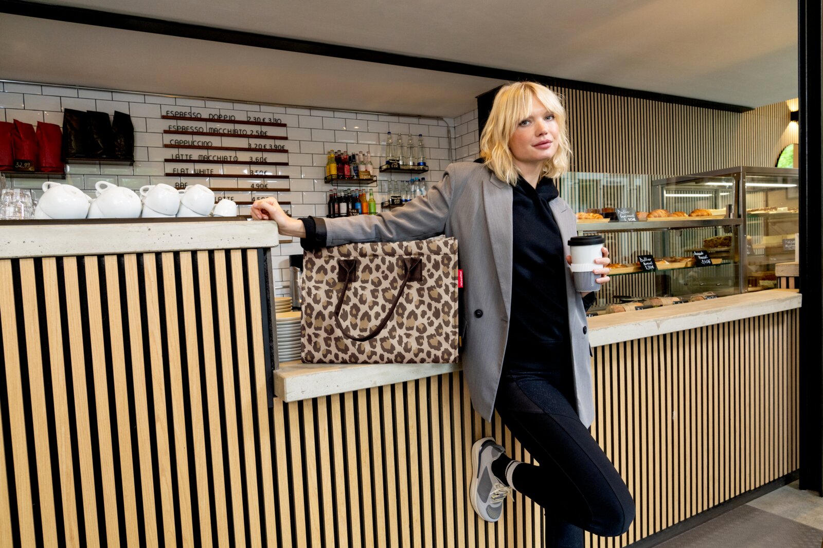 Frau lehnt an einer Theke in einem Café, hält einen Kaffeebecher und eine Tasche mit Leopardenmuster, seitliche Perspektive.