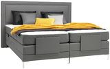 Graues Boxspringbett mit gestepptem Kopfteil, dekoriert mit schwarzen, grauen und gelben Kissen sowie einer gelben Decke, aus der Frontalperspektive.