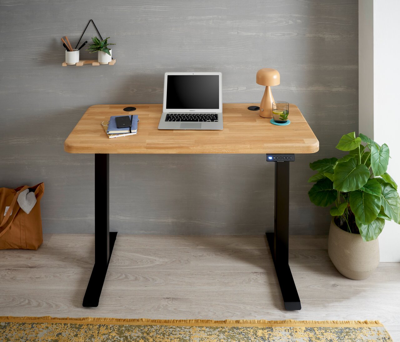 Moderner E-Schreibtisch mit Holzplatte und schwarzen Metallbeinen, frontal fotografiert. Auf dem Tisch stehen ein Laptop, eine Lampe und ein Glas. Eine Pflanze steht rechts daneben.