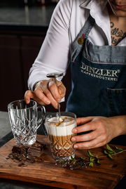 Nahaufnahme eines Barista, der einen Kaffeebecher mit Milchschaum zubereitet, neben einem leeren, kunstvoll geschliffenen Glasbecher auf einem Holzbrett mit Schokoladenstücken und Kräutern.