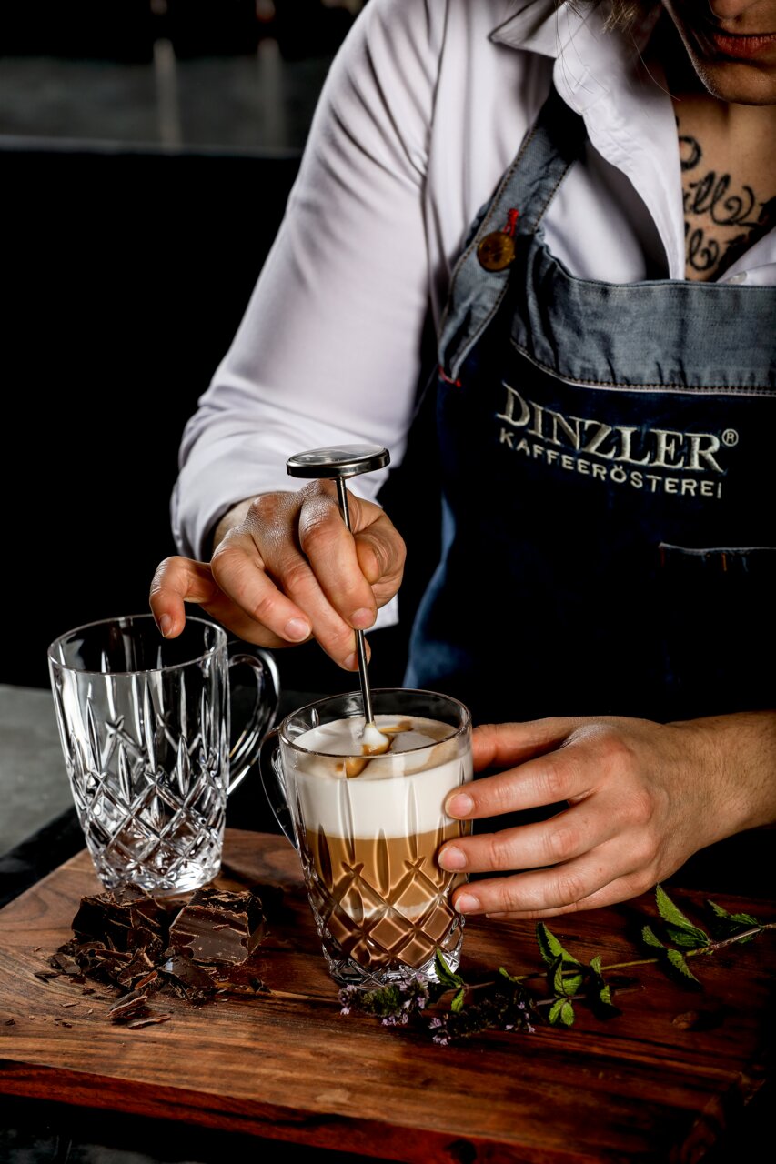 Nahaufnahme eines Barista, der einen Kaffeebecher mit Milchschaum zubereitet, neben einem leeren, kunstvoll geschliffenen Glasbecher auf einem Holzbrett mit Schokoladenstücken und Kräutern.