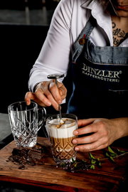 Nahaufnahme eines Barista, der einen Kaffeebecher mit Milchschaum zubereitet, neben einem leeren, kunstvoll geschliffenen Glasbecher auf einem Holzbrett mit Schokoladenstücken und Kräutern.