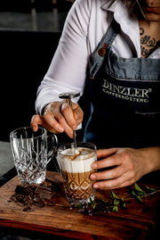 Nahaufnahme eines Barista, der einen Kaffeebecher mit Milchschaum zubereitet, neben einem leeren, kunstvoll geschliffenen Glasbecher auf einem Holzbrett mit Schokoladenstücken und Kräutern.