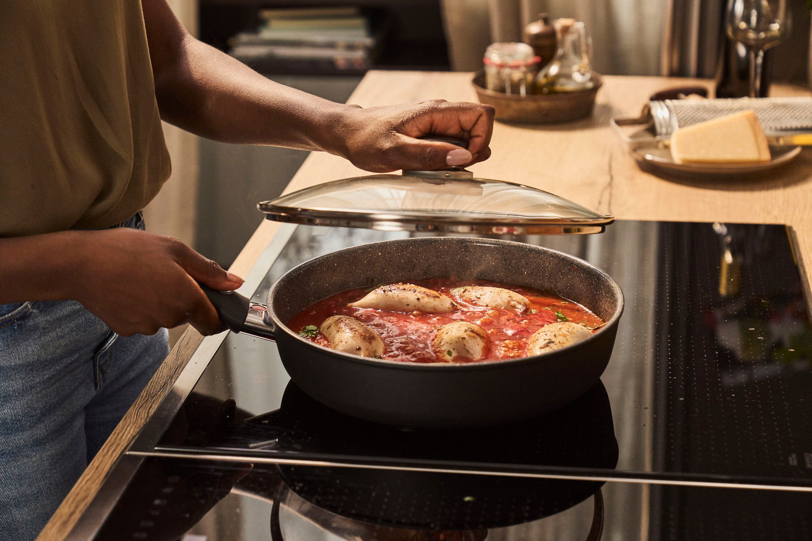 Person kocht in einer Schmorpfanne mit Glasdeckel auf einem Herd, seitliche Perspektive.