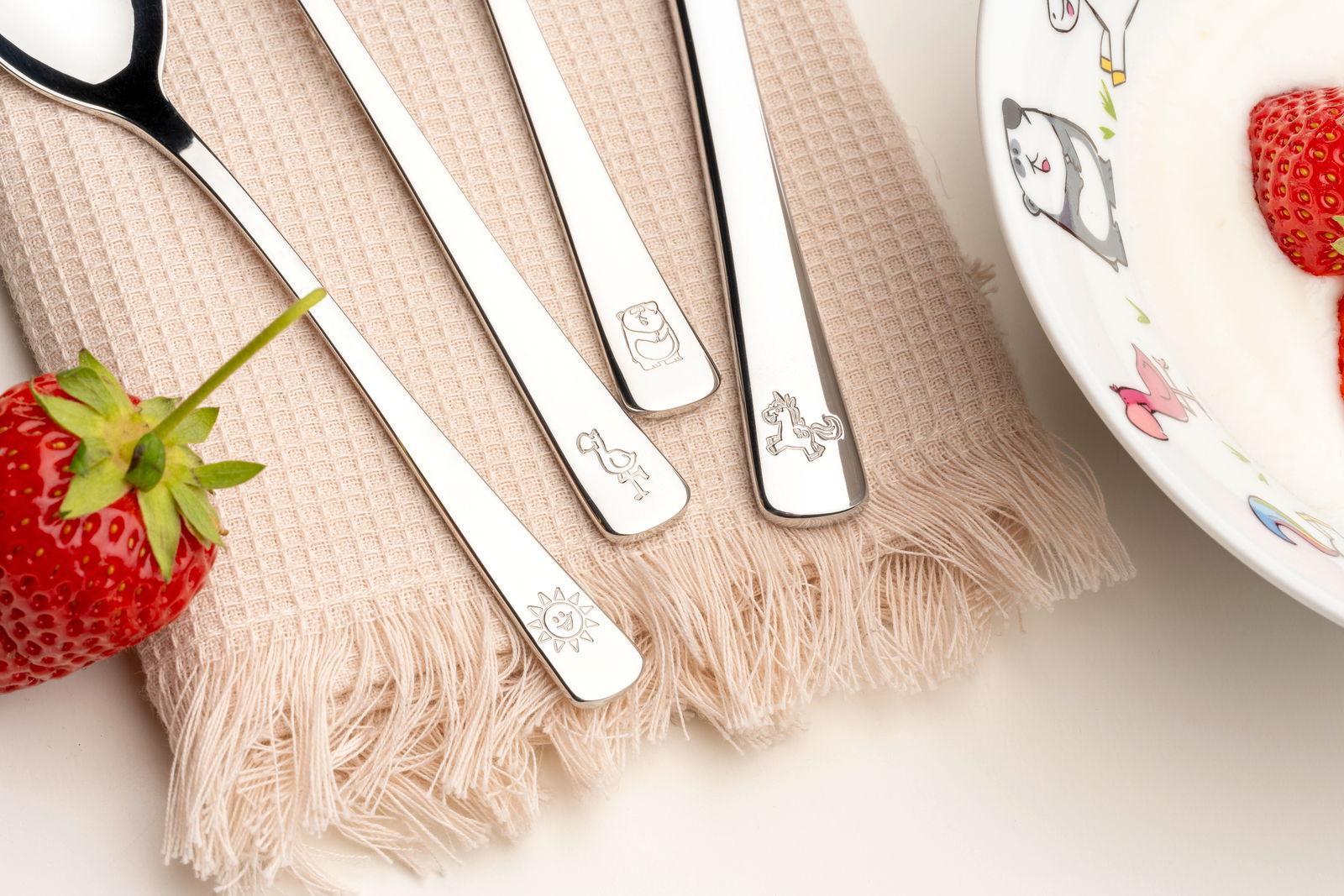 Leonardo Kinderbesteck 4-tlg. BAMBINI Kinderbesteck-Set mit vier Teilen, verziert mit Tiermotiven, auf einem beigen Tuch neben einer Erdbeere und einer Schale mit Joghurt. Draufsicht.