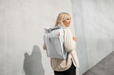 Frau mit einem hellgrauen urbanen Rolltop-Rucksack auf dem Rücken, seitliche Perspektive, vor einer Betonwand